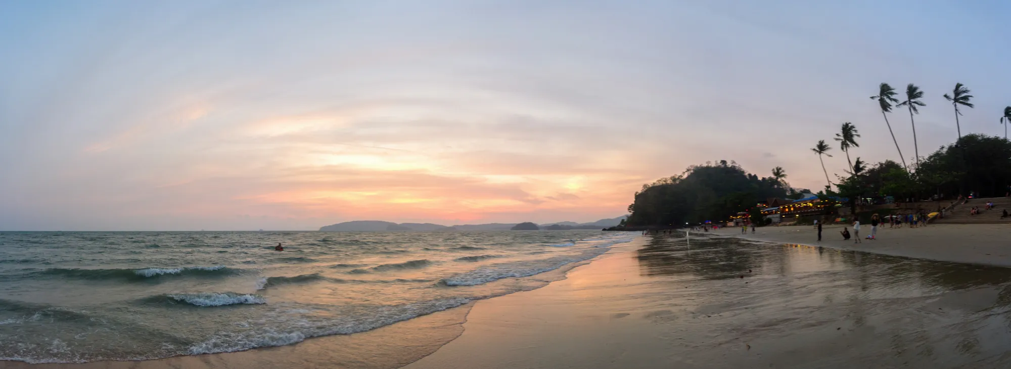 Ao Nang beach at sunset, with a pink sky, small waves on the sand and palm trees on the far shore