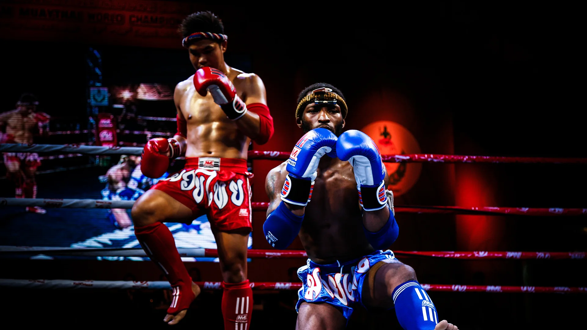 Two Muay Thai fighters performing the ram muay in a ring under stage lights, one standing on a raised knee in red gloves and one kneeling in blue gloves