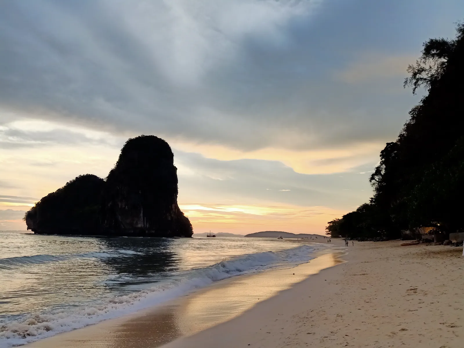 A limestone rock rising from the sea at Phra Nang Cave Beach at sunset, with wet sand and a dark cliff on the right