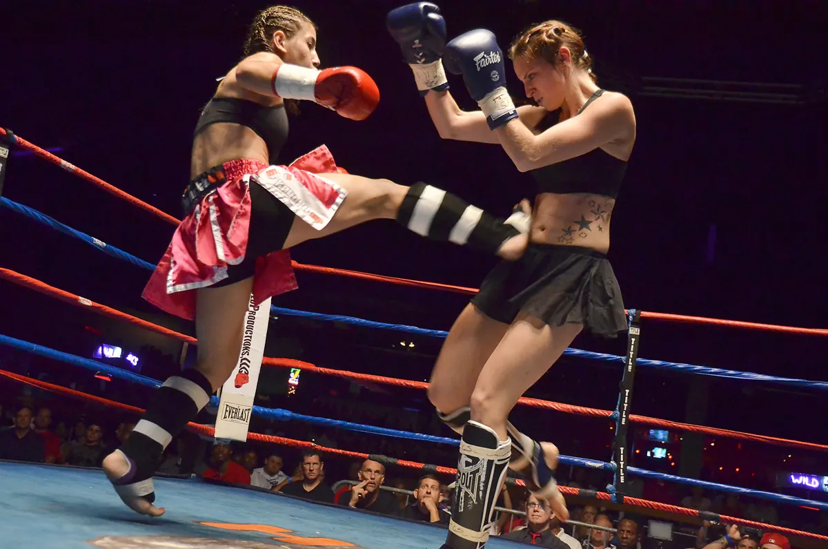 Two women fighters in a ring, one throwing a teep push kick into the other's guard while the crowd watches from ringside